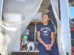 Frau sitzt im Eingang eines Wohnmobils und schaut in die Ferne, neben ihr liegt ein Welpe aus Griechenland
