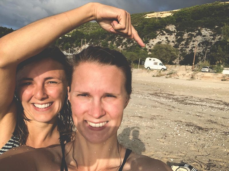 Zwei Frauen am Strand in Griechenland. die eine Frau hebt den Arm und zeigt auf den weißen Mercedes Sprinter, der im Hintergrund geparkt ist. Es ist ein ruhiger Ort um in Griechenland freistehen zu können