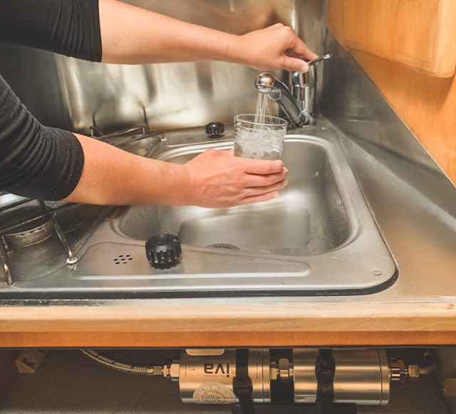 Ein Wasserglas wird unter einem Wasserhahn in einem Wohnmobil gehalten und mit Wasser befüllt. In der Schublade darunter sieht man zwei riva Waserfilter eingebaut, die das Wasser aus dem Tank vor der Entnahme filtern.