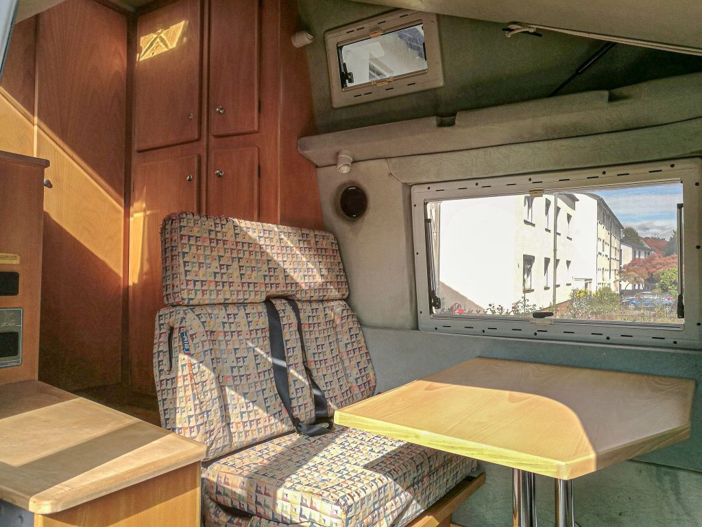 Das Bild zeigt das Innere eines Wohnmobils, das eine Erneuerung der Innenverkleidung benötigt. Es ist ein eingebauter Tisch und eine Sitzbank mit gemusterten, alten Polstern zu sehen. Hölzerne Schränke und ein Fenster, das einen Blick auf eine Wohngegend bietet, betonen das gemütliche, aber renovierungsbedürftige Design