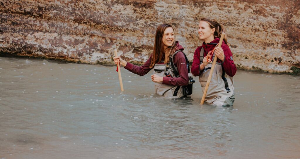 Zwei Frauen sind in Anglerhosen und halten Wanderstöcke in der Hand. Sie stehen bis zur Hüfte im Fluss im Zion Nationalpark und wandern The Narrows