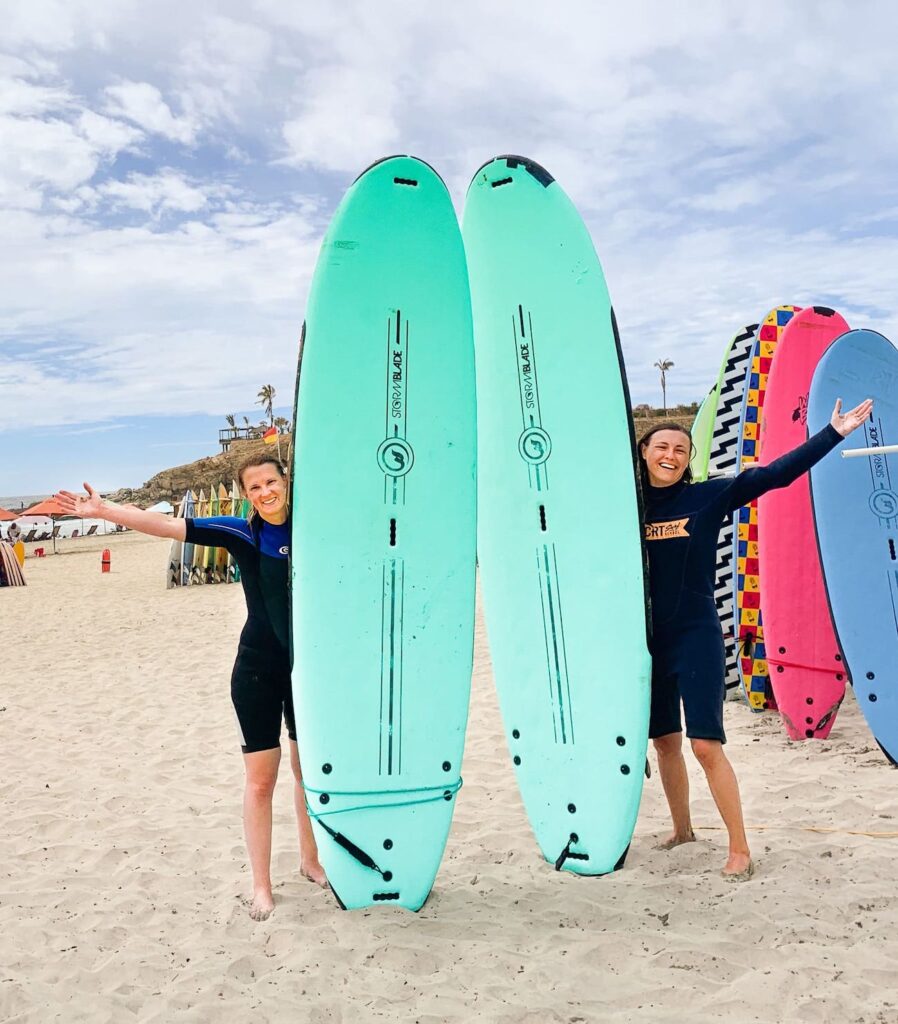 Zwei Frauen in Wetsuits stehen freudig neben zwei Surfbrettern in Mexiko. Mit der Young Travellers Auslandskrankenversicherung sind sie bestens versichert, auch beim Sport machen.