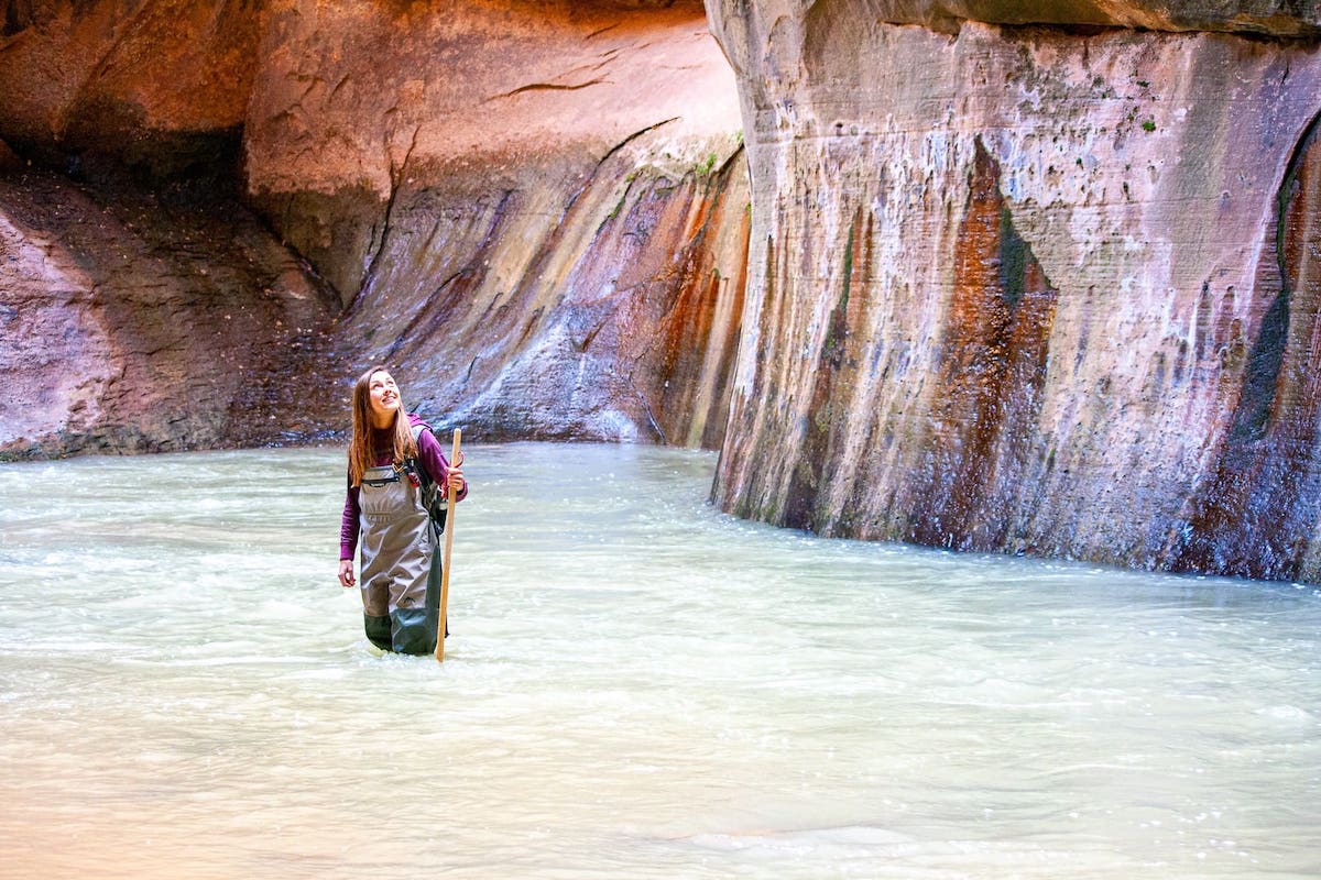 Dieses Bild zeigt eine Frau inmitten eines Abenteuers im Zion Nationalpark in den USA. Sie steht im Virgin River im Zion Nationalpark, hält ein Wanderstock in der Hand und schaut auf die Felswände, die sie umgeben. Der Fluss ist von hohen Klippen umgeben, und das Wasser ist kristallklar. Die Frau scheint einen Moment zu nutzen, um die natürliche Schönheit der Gegend zu schätzen, bevor sie ihre Wanderung fortsetzt.