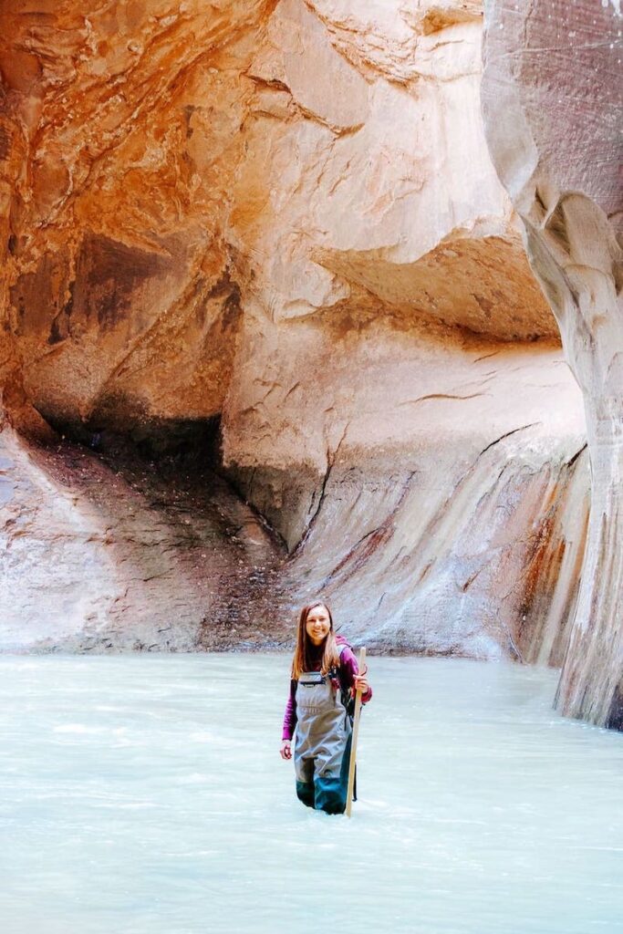 Eine Frau steht in einer Anglerhose und mit einem Wanderstock in der Hand im Virgin River im Zion Nationalpark. Sie wandert auf dem Wanderweg The Narrows, welcher stromaufwärts durch den Canyon führt 