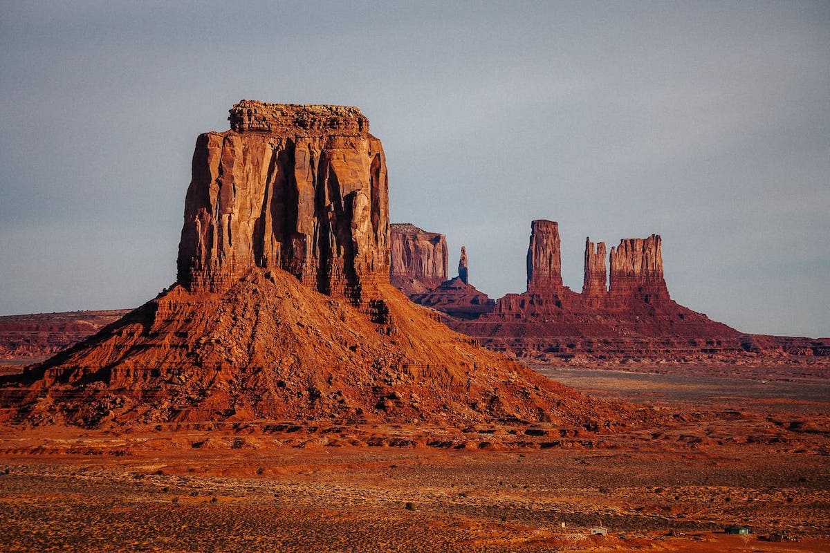 Dieses Bild zeigt die beeindruckenden Felsformation des Monument Valleys. Die Felsformation besteht aus mehreren hohen Felsen, die in einem tiefen Rotton schimmern. Der Himmel darüber ist diesig, ohne eine Wolke in Sicht. Dies ist das Monument Valley, ein Ort, den man bei einer Reise durch den amerikanischen Südwesten unbedingt besuchen sollte.