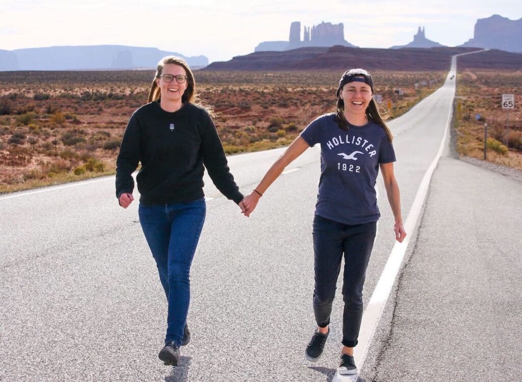 Zwei Frauen laufen auf der Straße vor dem Forest Gump Point. Im Hintergrund sieht man das Monument Valley