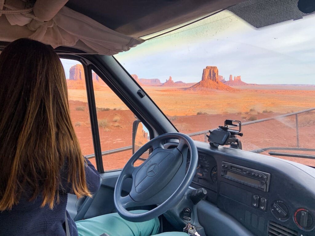Dieses Bild zeigt eine Frau, die im Wohnmobil durch das Monument Valley fährt. Die Frau schaut aus dem Fahrerfenster. Im Hintergrund erstrecken sich die charakteristischen roten Felsen des Monument Valley.
