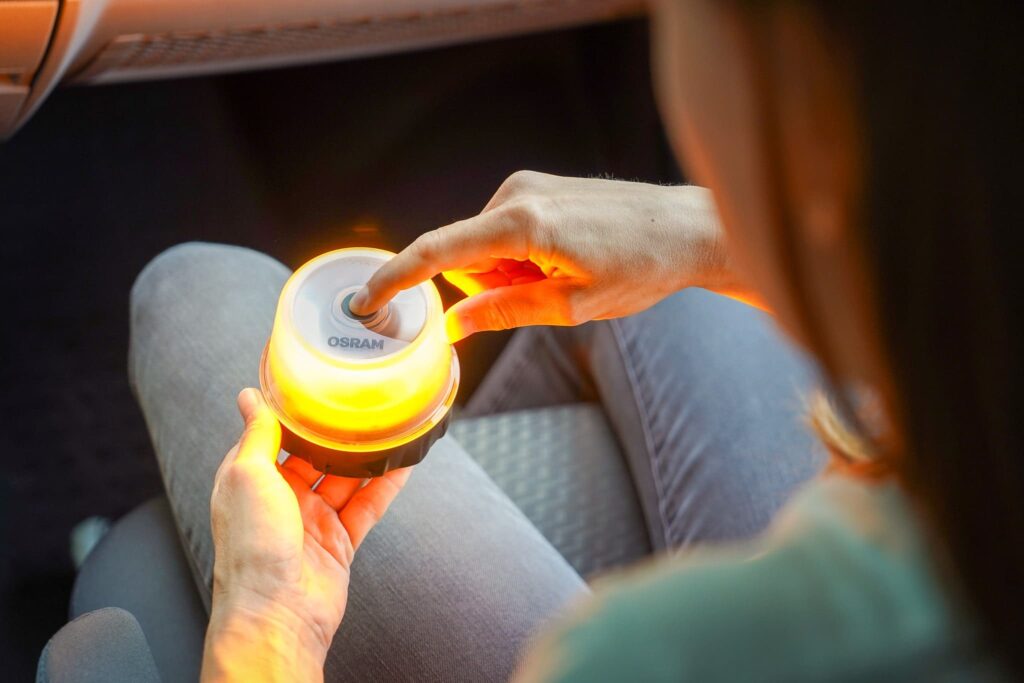 Eine Frau sitzt auf dem Beifahrersitz eines VW Bulli Wohnmobils und hält die Warnleuchte von OSRAM in der Hand. Ihr FInger ist auf dem Einschaltknopf und die Warnleuchte leuchtet hell orange.