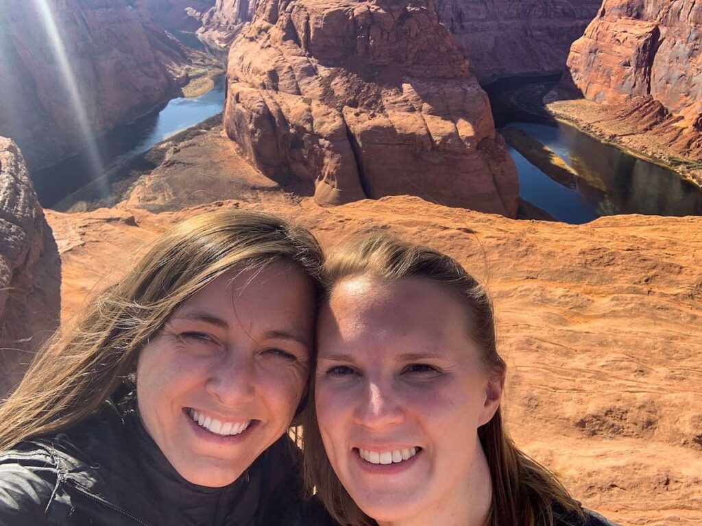 Zwei Frauen lächeln in die Kamera, im Hintergrund ist Horseshoe Bend in Arizona zu sehen. Die Sonne steht hoch am Himmel