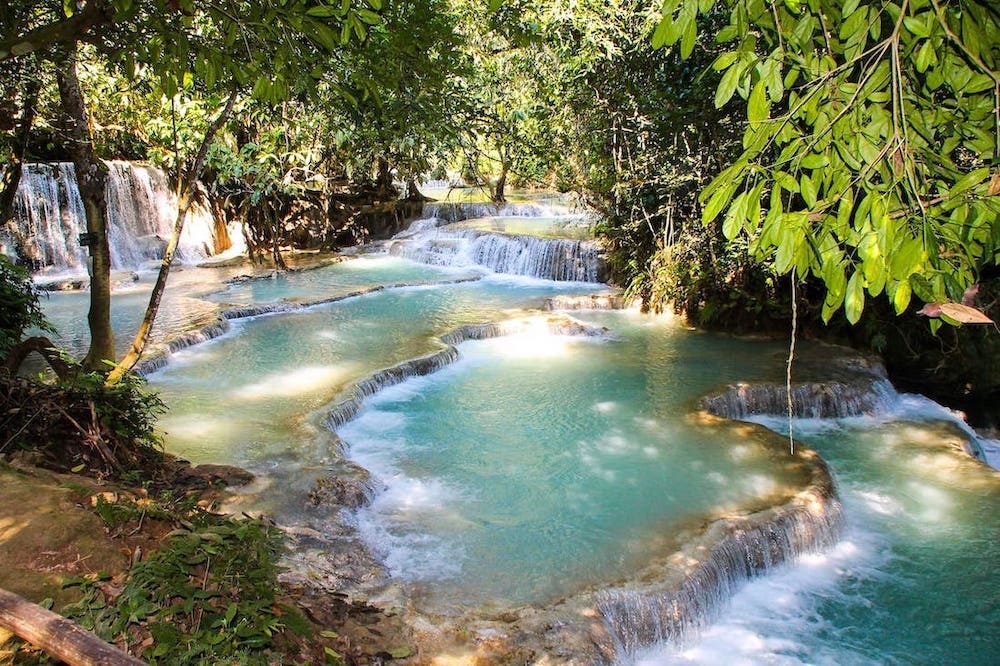 Das Bild zeigt ein türkisfarbenes Wasserbecken der Kuang Si Wasserfälle im Dschungel. Die Kuang Si Wasserfälle sind eine Sehenswürdigkeit in der Nähe von Luang Prabang