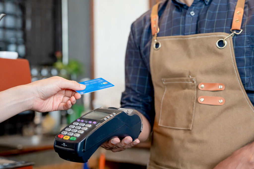 Eine Person bezahlt kontaktlos mit einer Kreditkarte an einem Kartenterminal in einem Café oder Geschäft. Der Kassierer, der eine braune Schürze mit Lederriemen trägt, hält das Zahlungsterminal bereit. Die Szene zeigt eine moderne und schnelle Zahlungsmethode per NFC-Technologie.